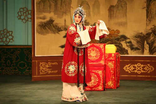 京剧《穆桂英挂帅》剧照:穆桂英着团花红蟒、持手帕立于桌案前(国家京剧院官网剧目页所载)