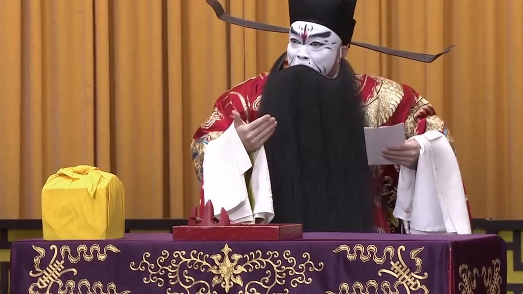 京剧《群英会·借东风·华容道》第四场演出照：红蟒、白脸黑满髯的净角立于桌案后（央视《CCTV空中剧院》）